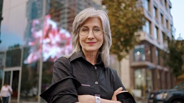 Portrait of middle age woman. Smiling senior woman with gray hair and glasses in stylish clothes. City street on blurred background. Active lady on retirement. Slow motion