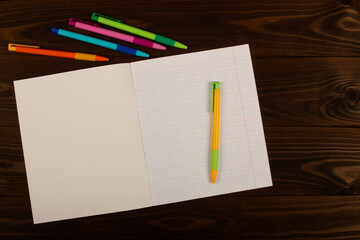 School notebook with oblique lines, multicolored pens on the table brown wooden background. Blank sheet of paper in notebook on the desk. Back to school. School supplies. Top view. Copy space for text