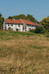 Meadow with a historic white half-timbered farm and trees in a hilly landscape under a blue sky on a sunny summer day.