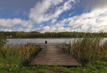Obraz premium Landscape with wooden long jetty with chair for fishing, lake, forest on horizon and cloudy blue sky in summer.