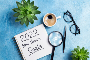 New year goals 2022 on desk. 2022 goals with notebook, coffee cup and eyeglasses on blue background. Resolutions, plan, strategy, action, idea concept