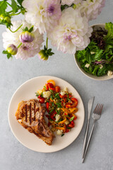 Delicious hearty dinner in plates on the table and a bouquet of beautiful autumn flowers, fried pork entrecote on the bone with stewed vegetables and fresh salad with mozzarella