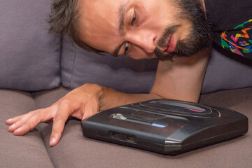 A bearded middle-aged young man in a T-shirt lay down on a sofa with a 16-bit retro video game...