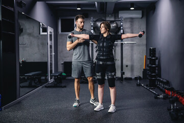 Fototapeta na wymiar A revolution in training, a new technological approach to training. A male instructor assists the women in a special suit for EMS technology while lifting dumbbells with both hands in a modern gym