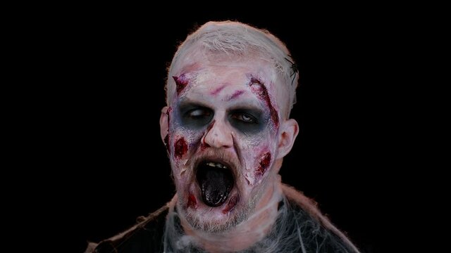 Frightening man with Halloween zombie bloody wounded makeup, trying to scare, showing tongue. Convulsions. Sinister undead guy isolated on studio black background. Voodoo rituals. Fashion body art