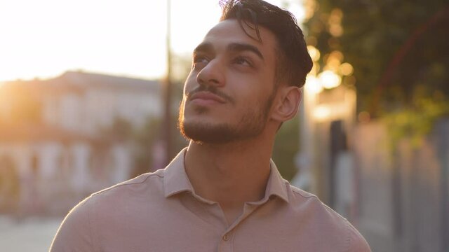 Close-up male happy inspired bearded face. Portrait arabic hispanic business man young adult pensive guy stands in city on street background of sunset sun dreaming thinking enjoying rays in summer
