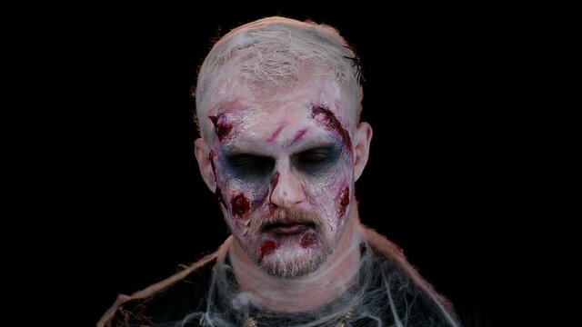 Portrait of sinister man in carnival costume of Halloween crazy zombie with bloody wounded scars face trying to scare isolated on black room. Horror theme of cosplay wounded undead, beast, monster