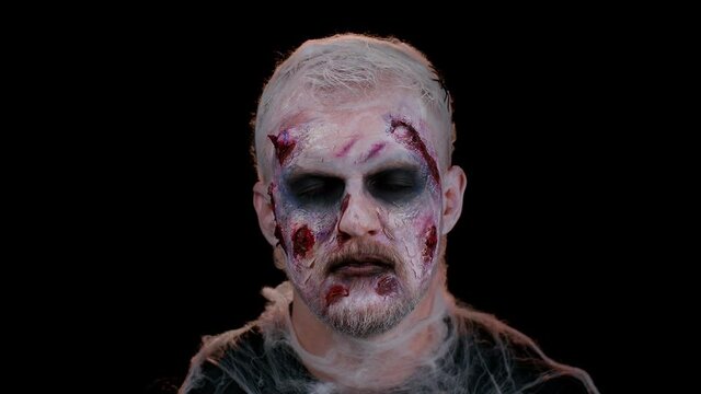 Unexpected appearance of sinister man with horrible scary Halloween zombie makeup making faces, looking ominous at camera, trying to scare. Dead guy with wounded bloody scars face. Black background