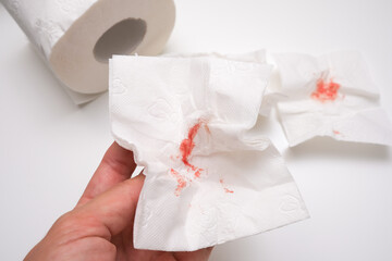 Hand holding used sheet of bloody toilet paper and a roll on white background, hemorrhoids and rectal bleeding concept