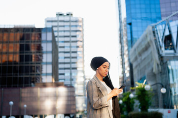 Side view of an adult muslim woman standing in a urban area using her phone