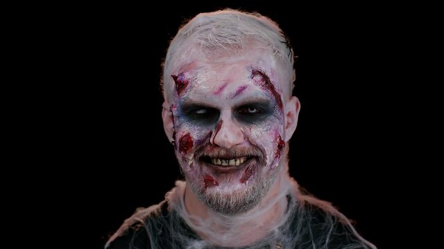 Playful sinister man Halloween crazy zombie with bloody wounded scars face blinking eye, looking at camera with toothy smile, winking and flirting, expressing optimism. Cosplay wounded undead monster