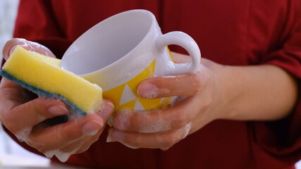 Close-up of a man washing a yellow mug.The man holds the dishes and washes them with a yellow soapy sponge. Dishes covered with soapy foam. Daily cleaning service. Washing dishes after eating. 