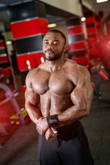 Fototapeta na wymiar Portrait of african american male fitness trainer. Bodybuilder showing his biceps and looking at the side of the gym background