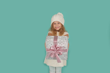 a beautiful happy little blue-eyed blonde girl in a knitted winter hat and a knitted white dress holds a gift box