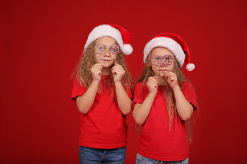 beautiful happy little blue-eyed blonde girls sisters in red T-shirts on their heads santa Claus hat on a red background holding a gift box the new year is holding the numbers 2022