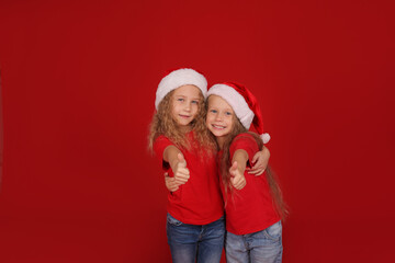 beautiful happy little blue-eyed girls blonde sisters in red t-shirts with santa Claus hat on their head on a red background hugging
