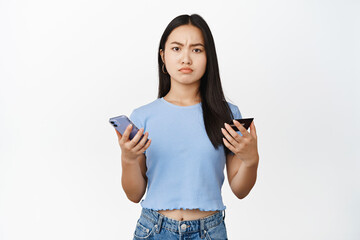 Concerned asian girl holding mobile phone and credit card, frowning and looking skeptical, standing in t-shirt over white background