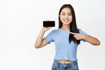 Fototapeta premium Cheerful asian girl pointing finger at horizontal smartphone screen, smiling while showing mobile application or store, white background