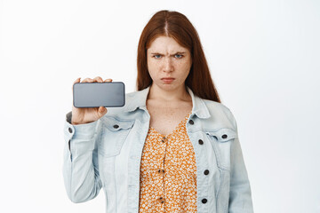 Angry teen redhead girl looking with disdain, showing mobile phone screen, standing upset against...