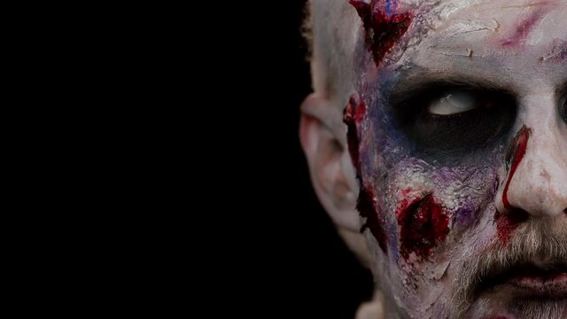 Close-up face of sinister man with horrible scary Halloween zombie makeup making faces, looking ominous at camera, trying to scare. Dead guy with wounded bloody scars face isolated on black background