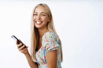 Naklejka premium Smiling blond girl holding smartphone, posing with mobile phone against white background