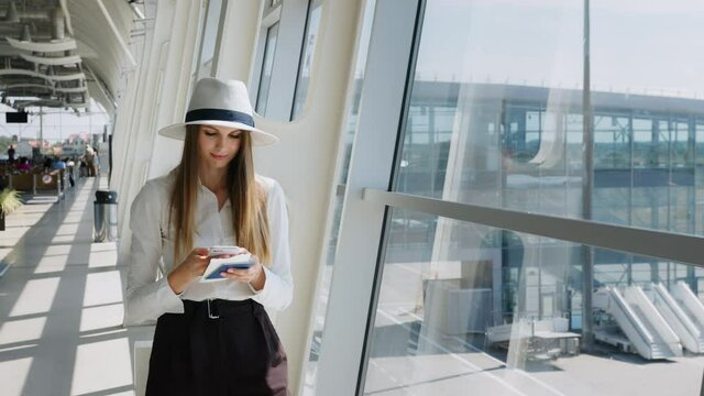 Elegant beautiful girl holding travel tickets and using a phone, happy girl looking at the camera in the airport terminal. Fashion woman portrait. Airport terminal. Business trip