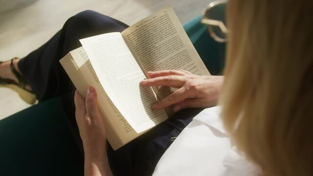 Elderly woman reading book, sitting on sofa in living room. Female pensioner studying literature, flipping through pages, spare free time at home during pension. 