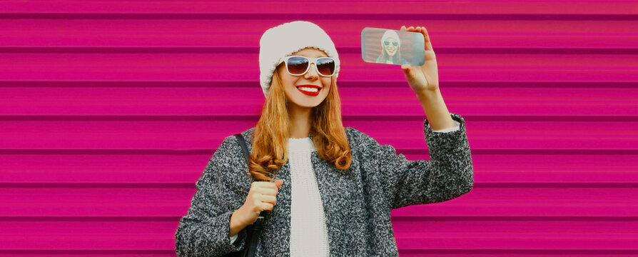 Portrait of happy young smiling woman taking a selfie by smartphone wearing a knitted white hat on colorful pink background
