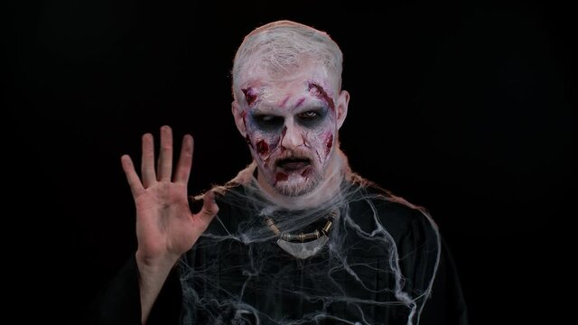 Creepy scary man with bloody scars face, Halloween zombie make-up. Scary wounded undead guy waves hand palm in hello gesture welcomes someone with hospitable expression expresses positive emotions