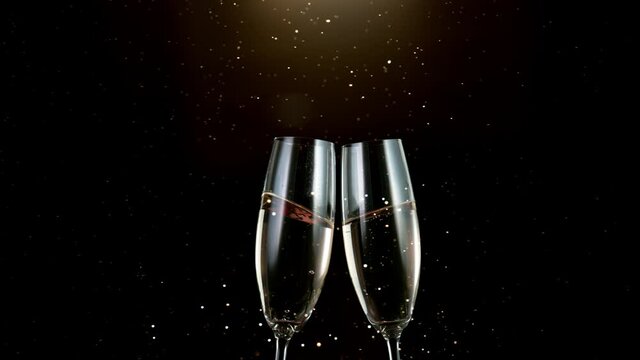 Super slow motion of toasting champagne flutes. Two glasses with sparkling champagne and golden particles falling, black background.