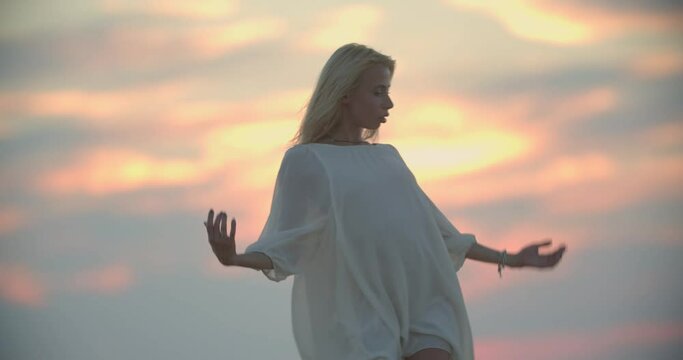 Beautiful blonde woman in white dress is dancing on the pier, sunset, 4k