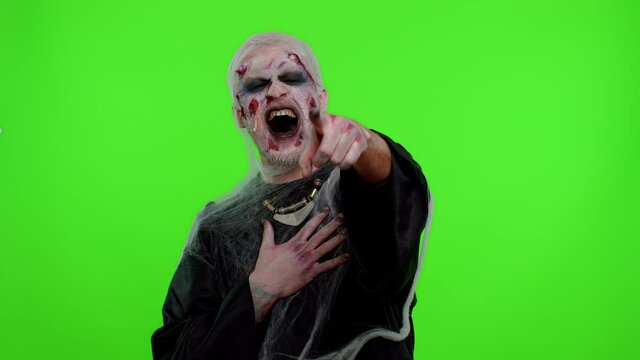 Sinister man in carnival costume of Halloween crazy zombie with bloody wounded scars face pointing finger to camera, laughing out loud, taunting making fun of ridiculous appearance, funny joke. Horror