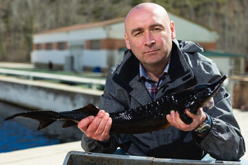 Confident fish farmer holding sturgeon grown in own fish husbandry..