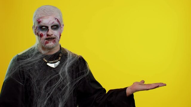 Creepy man with bloody scars face, Halloween stylish zombie make-up. Scary wounded undead guy showing thumbs up and pointing at right on blank space. Place for advertisement logo. Sinister monster