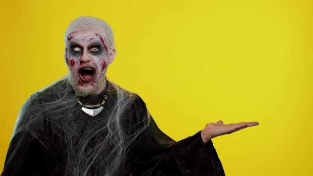 Zombie man with makeup with fake wounds scars showing thumbs up and pointing empty place, advertising area for commercial text logo, copy-space for goods promotion. Sinister Halloween ugly dead guy