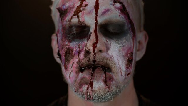 Close-up of sinister man with horrible scary Halloween zombie makeup making faces, looking ominous at camera, trying to scare, blood flows and drips on face. Dead guy with wounded bloody scars face