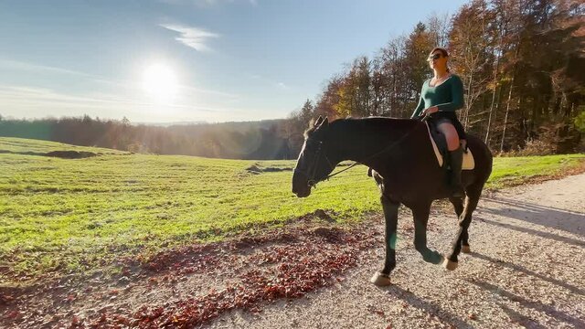 SLOW MOTION, LENS FLARE: Fit horseback rider guides her horse along a scenic route in rural Slovenia. Young woman rides her beautiful chestnut horse along a gravel path in autumn colored countryside.