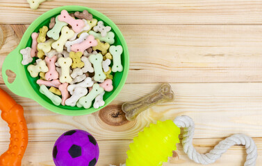 Different multicolored pet care accessories: green bowl, bones, balls, snacks on natural wooden background. Rubber and textile accessories for dogs. Top view, flat lay. Copy space	