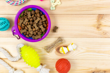 Different multicolored pet care accessories: violet bowl, bones, balls, snacks, toys on natural wooden background. Rubber and textile accessories for dogs. Top view, flat lay. Copy space	
