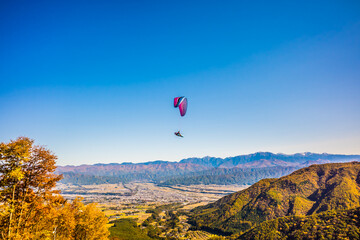 青空の下、紅葉の山を飛ぶパラグライダー
