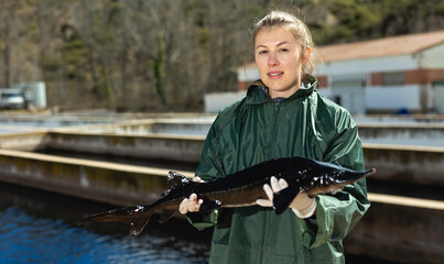 Proud young female owner of fish farm standing near pools with fresh sturgeons in hands..