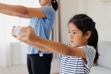 Asian young mother and her daughter doing stretching fitness exercise yoga and using the bottle of water to be dumbbells together at home in daily routine.
