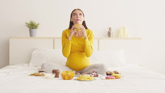 Food satisfaction. Young happy pregnant lady eating unhealthy hamburger with great pleasure, enjoying junk food delivery
