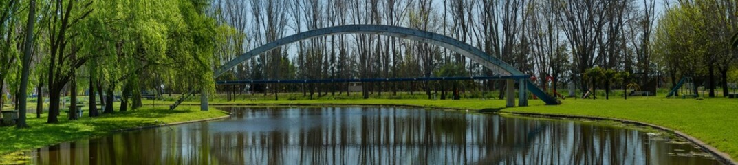 Panoramic view of an old concrete pedestrian bridge over a small lake in a park located in General...