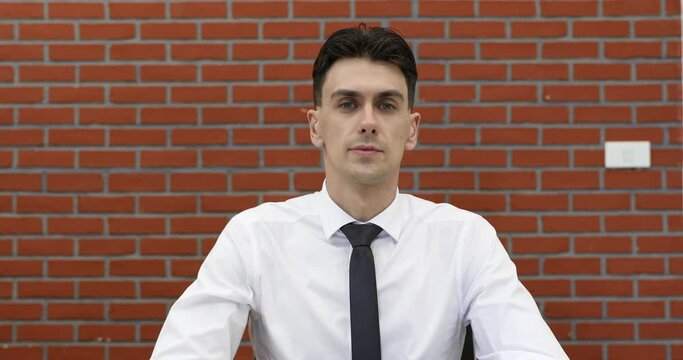 Young adult businessman in casual white shirt sitting in front of red brick wall listen to speaker in online conference and nod head with clear understanding.
