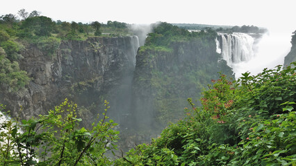 Fototapeta premium Victoria Falls streams descend into the gorge. The fog rises over the abyss. In the foreground, lush green vegetation, picturesque rocks. Zimbabw