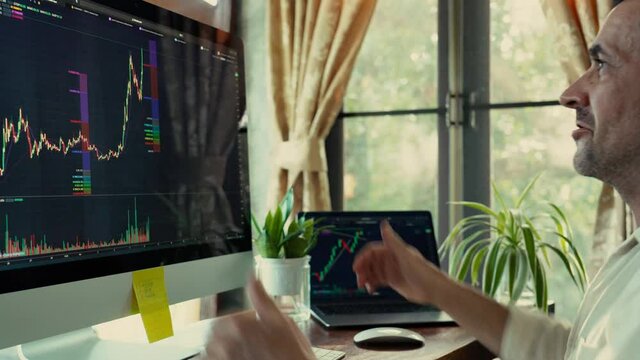 A man trader in a white shirt sits at a wooden table and uses desktop computer and laptop  at home