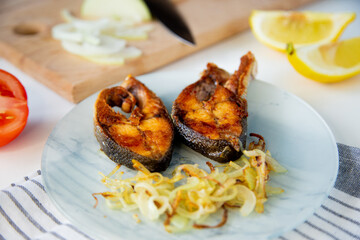 Fried fish with onions, lemon is on a plate, on a waffle towel, next to it is a cutting board with chopped onions on a white background. 