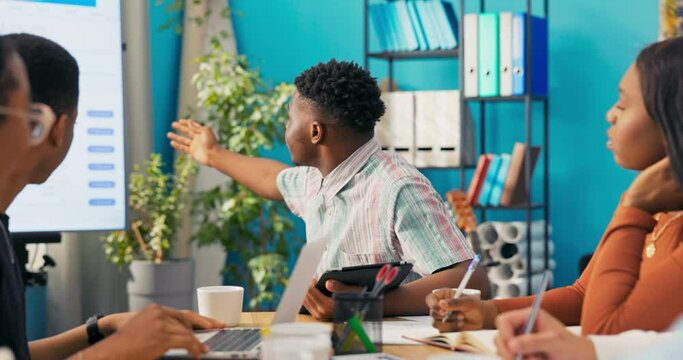 Dark skin man in striped collared shirt sits at large table in office with tablet looks at chart behind him on white interactive whiteboard asks colleague for interpretive details points with hand
