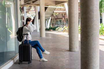 Traveling. Woman in a white long-sleeved shirt and jeans, wearing a mask. sit on the chair during travel with suitcase placed on her right hand and backpack